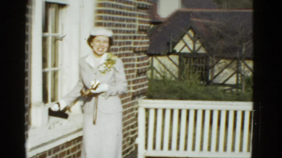 A smiling woman in a light suit and hat holds a camera beside a white porch railing in 1975 Boston footage.