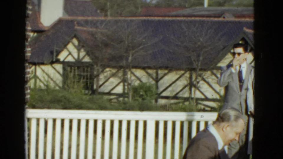 The frame shifts toward the porch railing and building exterior, with two adult men partly visible at the right edge.