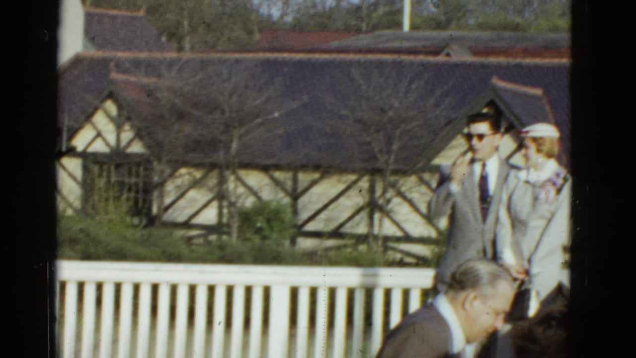 In 1975 Boston home-movie footage, a suited man in sunglasses stands with a woman in white beside a white railing and Tudor-style building.