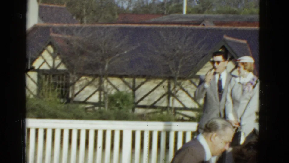 In 1975 Boston home-movie footage, a suited man in sunglasses stands with a woman in white beside a white railing and Tudor-style building.