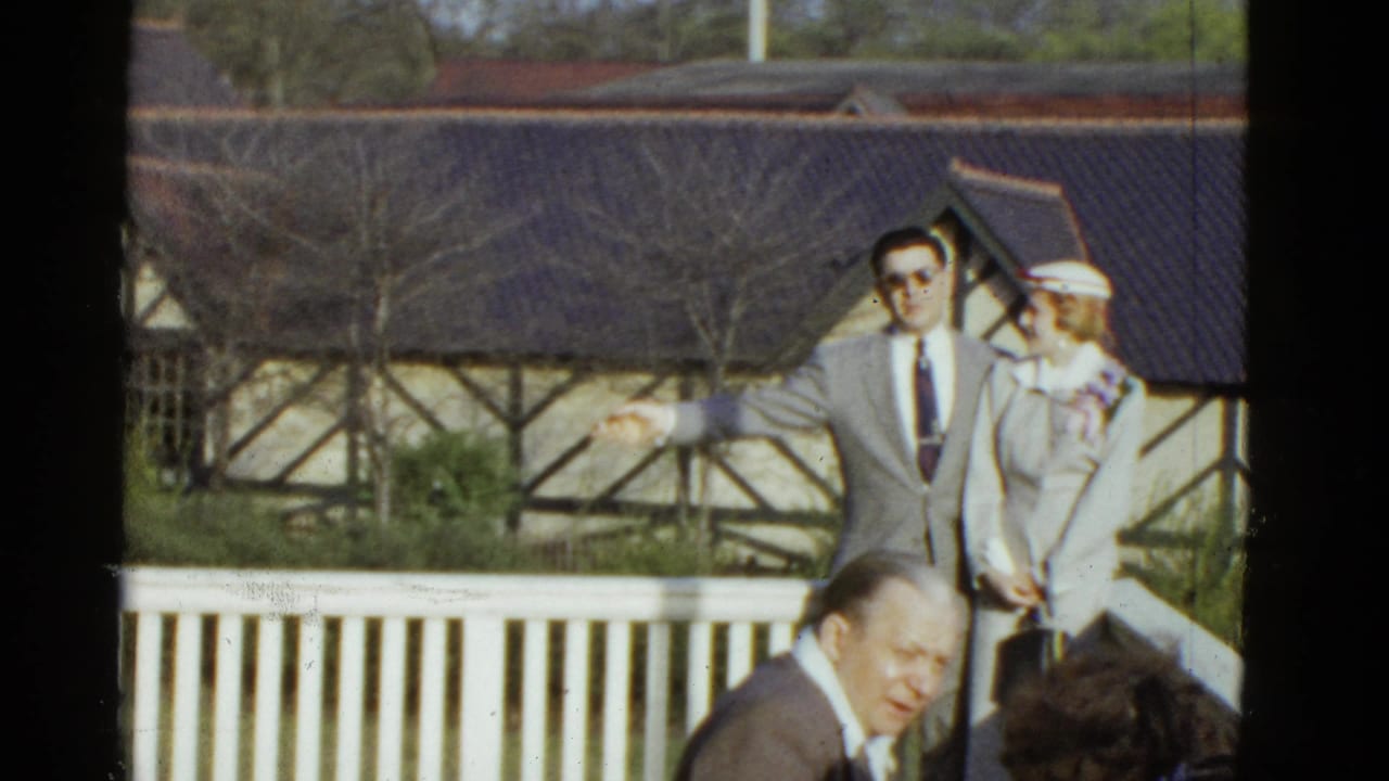 Suited man gestures beside a woman in white while guests sit in the foreground.