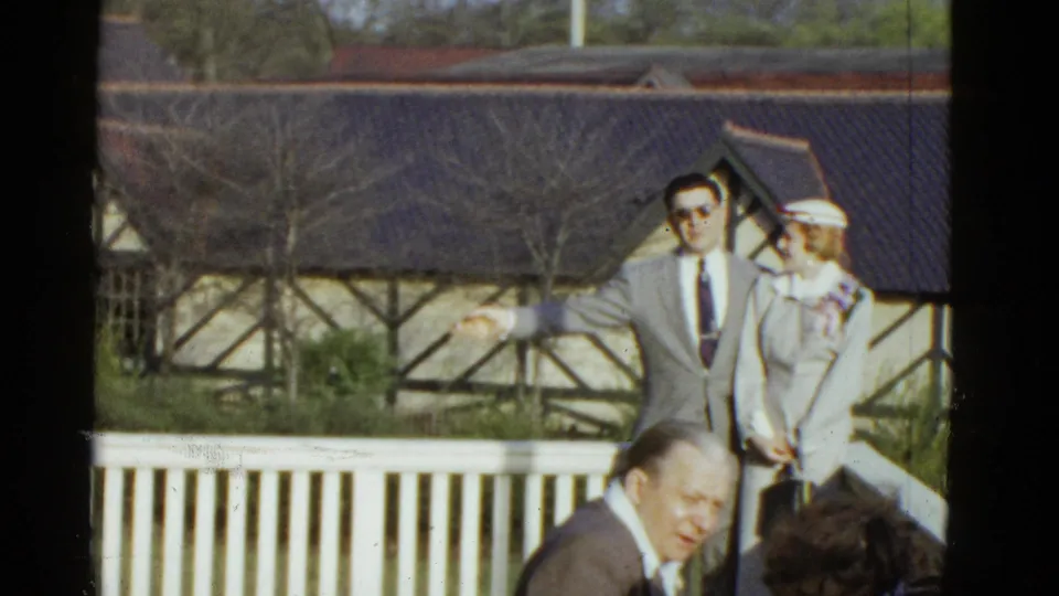 The suited man gestures outward while standing with a woman in white at an outdoor Boston gathering, with seated guests partly visible below.