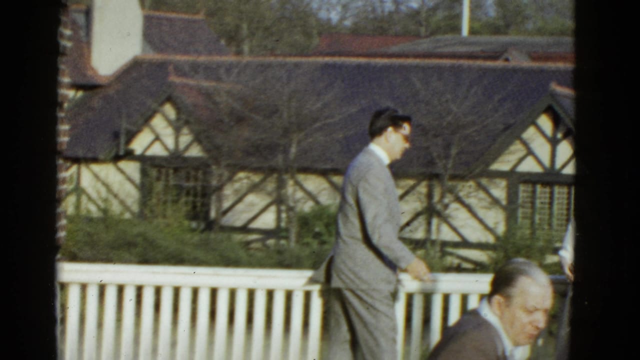 Man in a light gray suit walks beside a white railing with a Tudor-style building behind him.