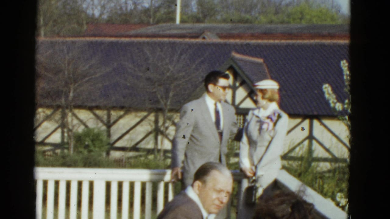 1975 Boston frame with a man in sunglasses and a woman in a white suit and hat near a porch railing and timber-framed building.