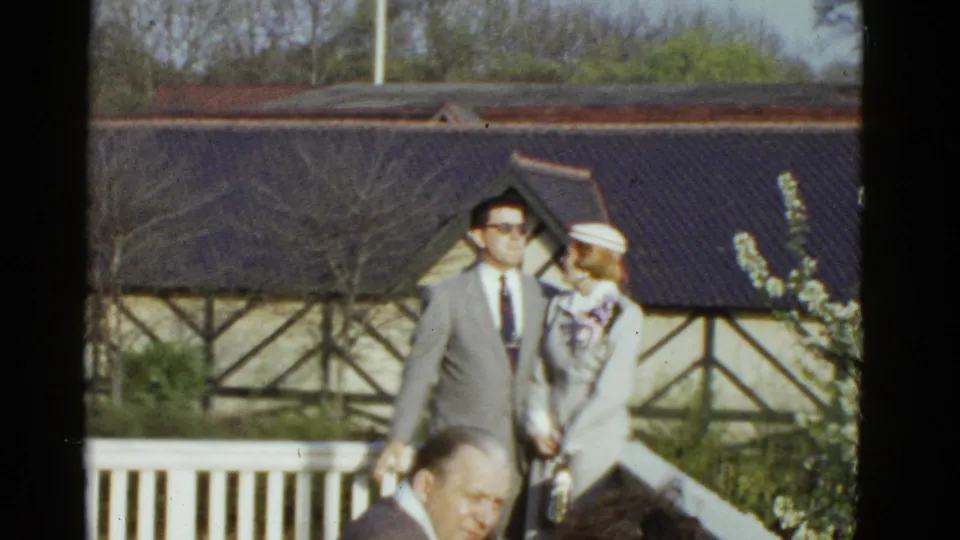 1975 Boston home-movie frame of a dressed man and woman walking on a sunny porch with a Tudor-style building behind them.