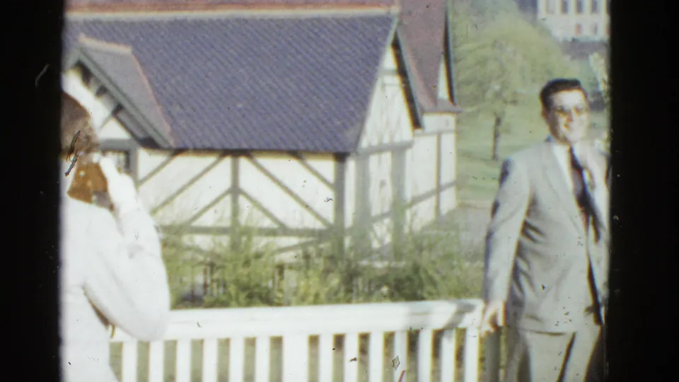 1975 Boston frame showing a suited man near the edge of a porch view, with a timber-framed building and railing behind him.