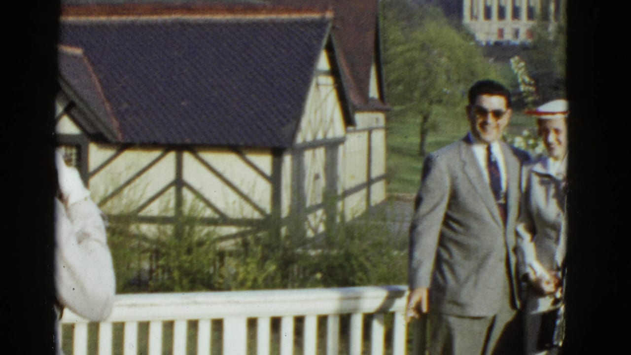 Man in a gray suit walks beside a woman near a white railing and Tudor-style building.