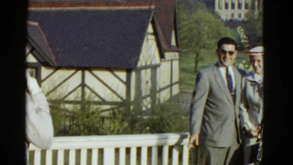 1975 Boston home-movie frame showing a suited man and woman near a white railing with a Tudor-style building behind them.