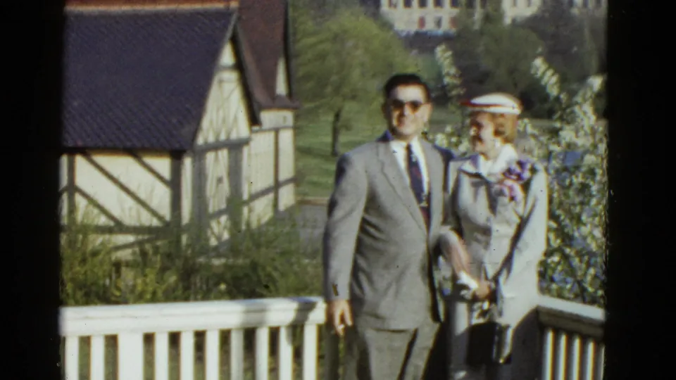 1975 Boston 8mm frame of a man in sunglasses and a woman in a hat near a white railing and Tudor-style structure.