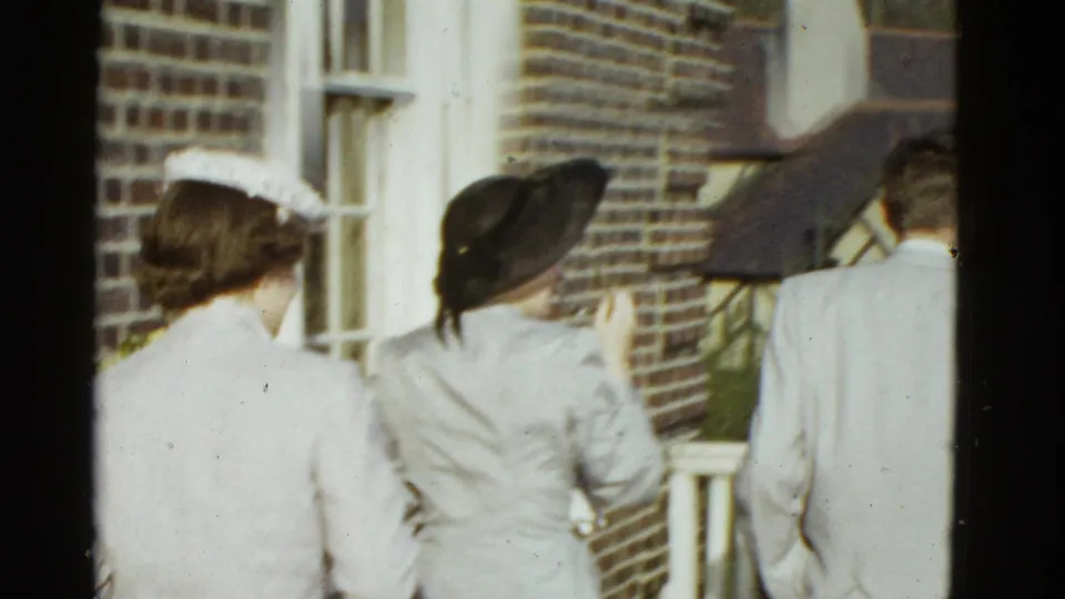 1975 Boston 8mm frame of adults in light formal clothing walking beside a brick building and white railing.