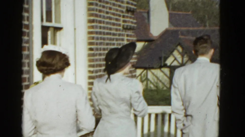 In 1975 Boston home-movie footage, two women in hats and a man in a light suit approach a balcony beside a brick exterior.