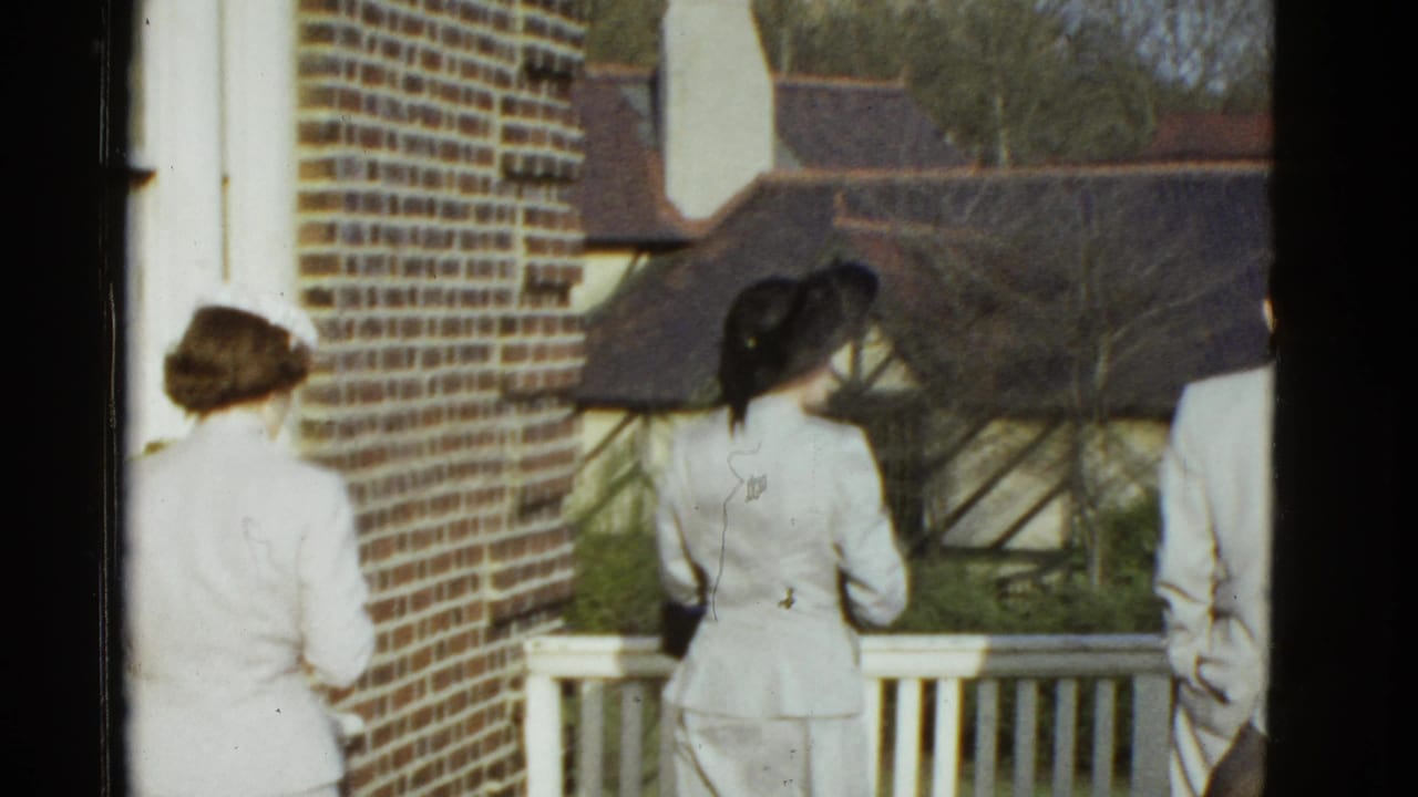 Two women in hats and a man in a light suit pause near a white railing in 1975 Boston home-movie footage.