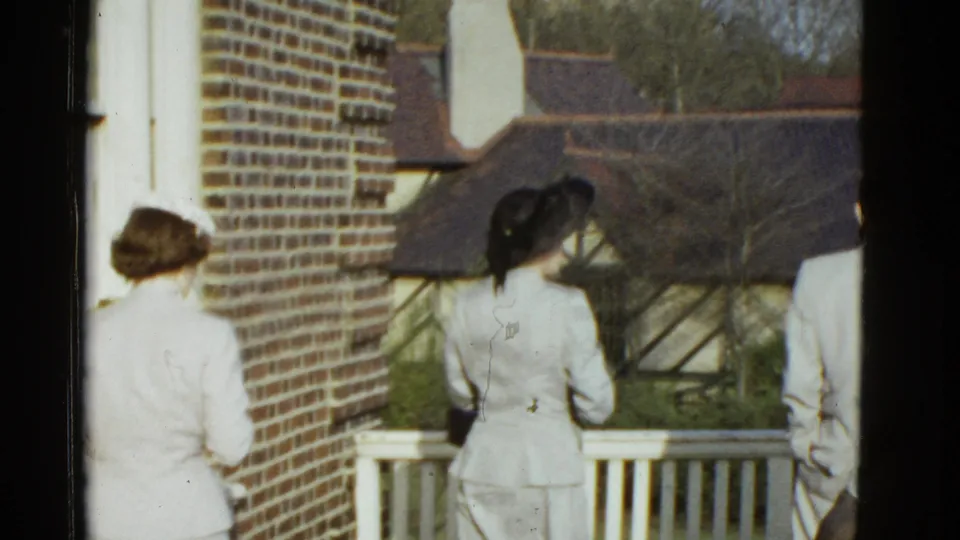 Two women in hats and a man in a light suit pause near a white railing in 1975 Boston home-movie footage.