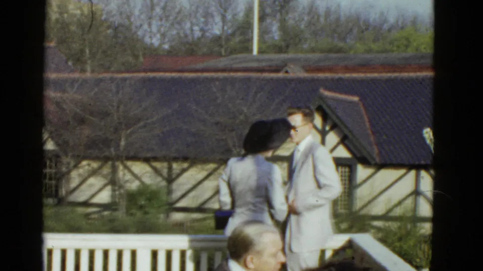 A man in a light suit turns toward a hatted woman beside a white balcony railing in 1975 Boston home-movie footage.