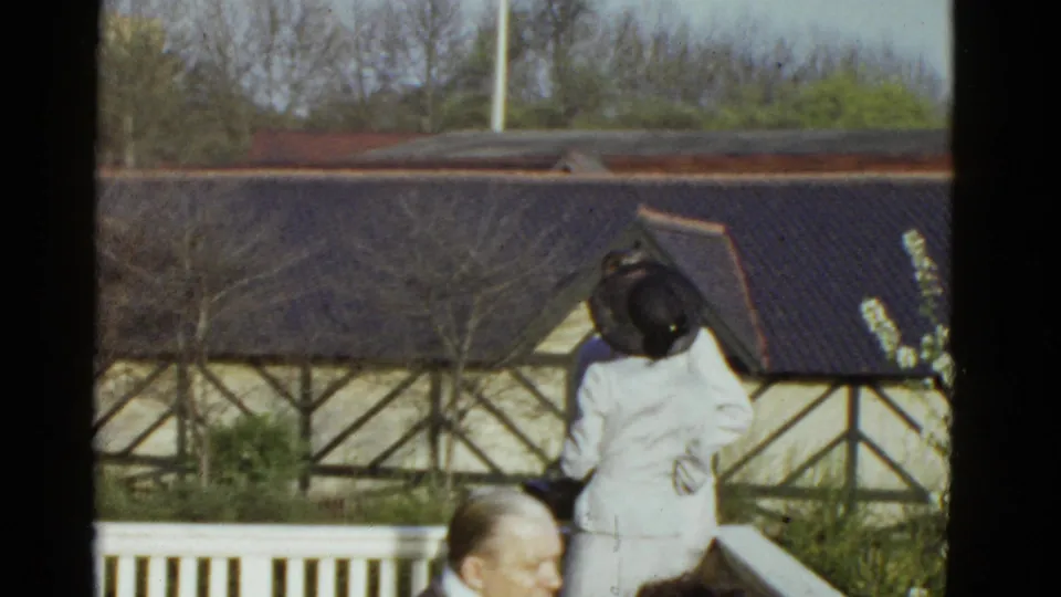 1975 Boston home-movie frame of a woman in a pale suit and dark hat standing by a porch railing with a seated man partly visible.