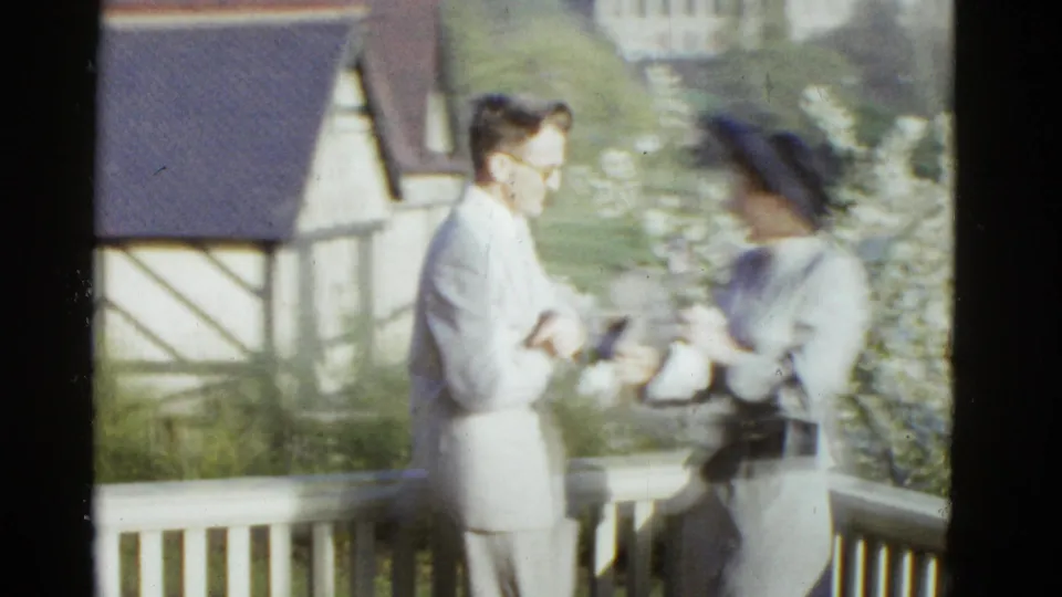 1975 Boston 8mm frame showing a well-dressed man and woman conversing on a porch with a half-timbered building behind them.