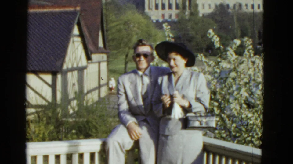 1975 Boston 8mm frame of a seated couple on a porch, with the man in sunglasses and the woman holding a handbag near flowering shrubs.