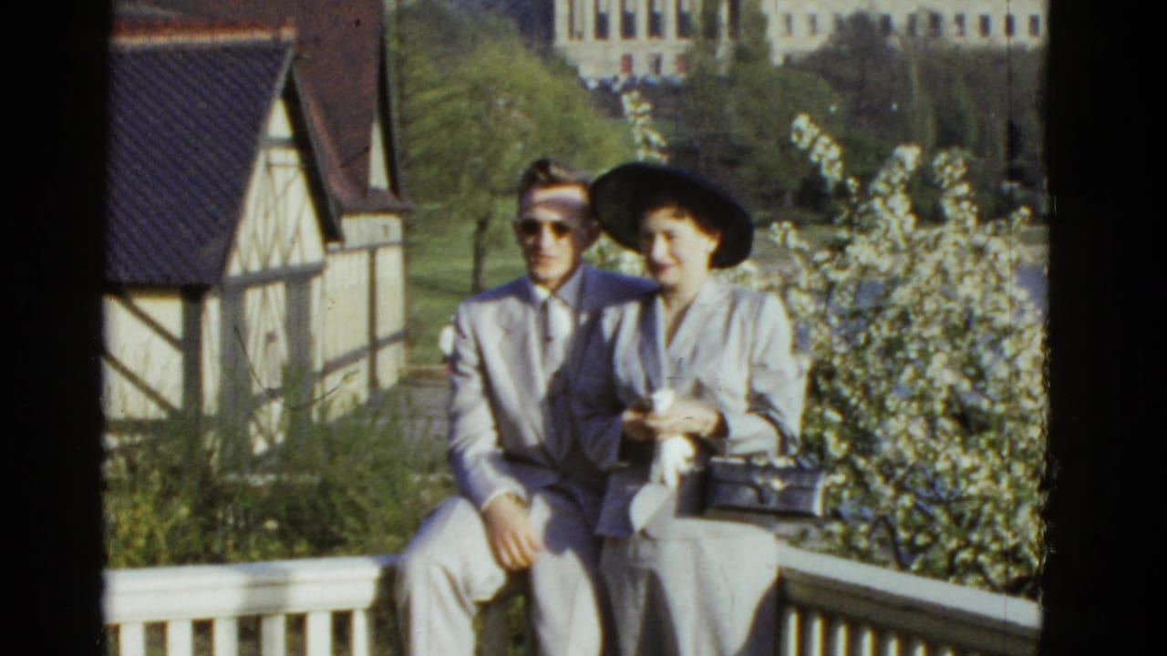 The couple remains seated on the balcony, with the woman holding a small purse.
