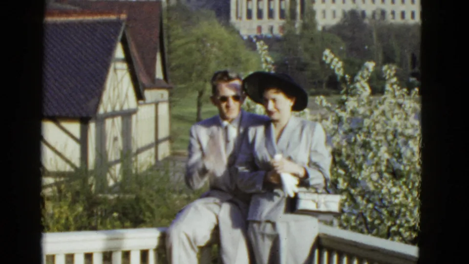 The 1970s home-movie frame shows the couple seated shoulder to shoulder, with a Tudor-style structure and springlike shrubs behind them.