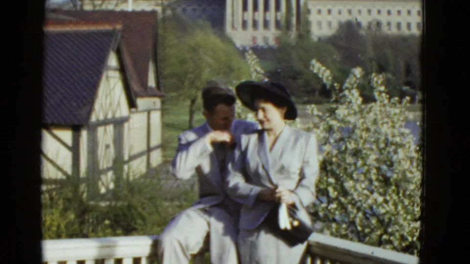 In 1975 Boston home-movie footage, a man and a woman in dress clothes sit close together on a balcony near flowering trees.