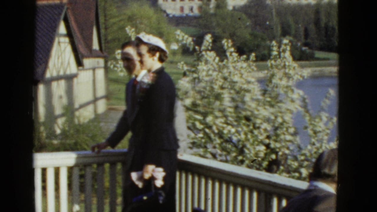 A dressed man and woman walk along a balcony beside blooming trees and a white rail.