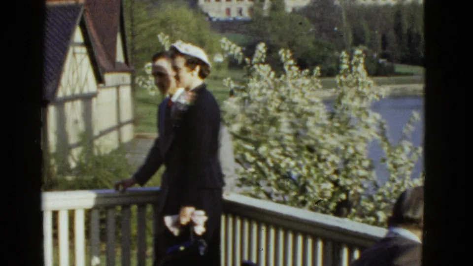 A dressed couple walks together on a Boston balcony, with the woman wearing a light hat and dark outfit in 1975 home-movie footage.