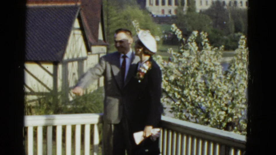 A suited man gestures while standing with a woman in a hat and dark coat on a balcony in 1975 Boston home-movie footage.