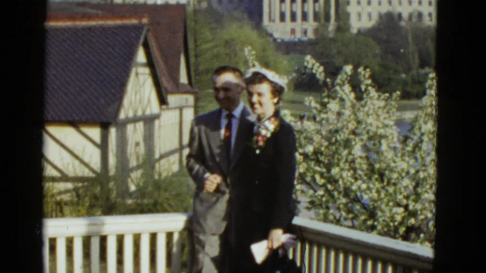 A dressed couple stands arm in arm on a Boston balcony, smiling toward the camera amid flowering trees in 1975 8mm footage.