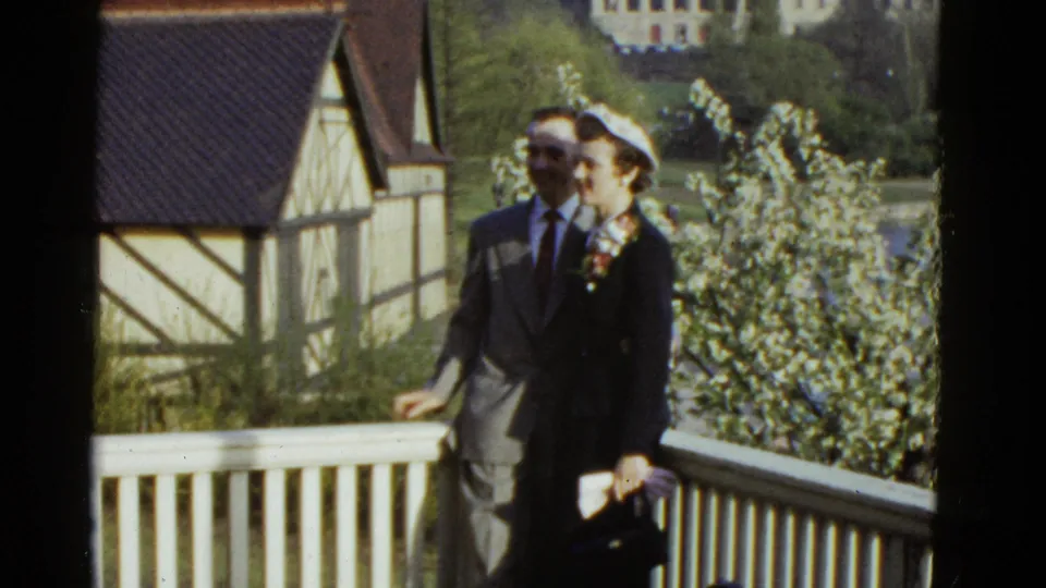 The couple remains posed on the balcony, with the man in a gray suit and the woman holding a small purse in 1975 Boston footage.