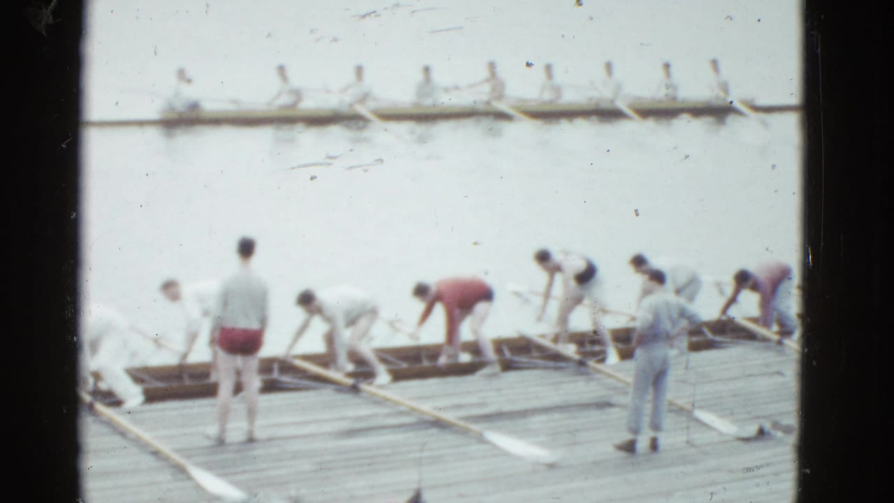 At a Boston rowing site in 1975 home-movie footage, crew members prepare rowing shells on a dock as another boat moves across calm water.