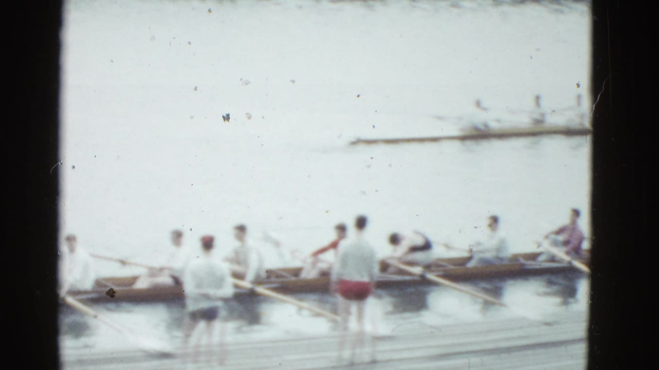 1975 Boston 8mm frame showing rowers in a long shell near a wooden dock, with standing figures watching or assisting.