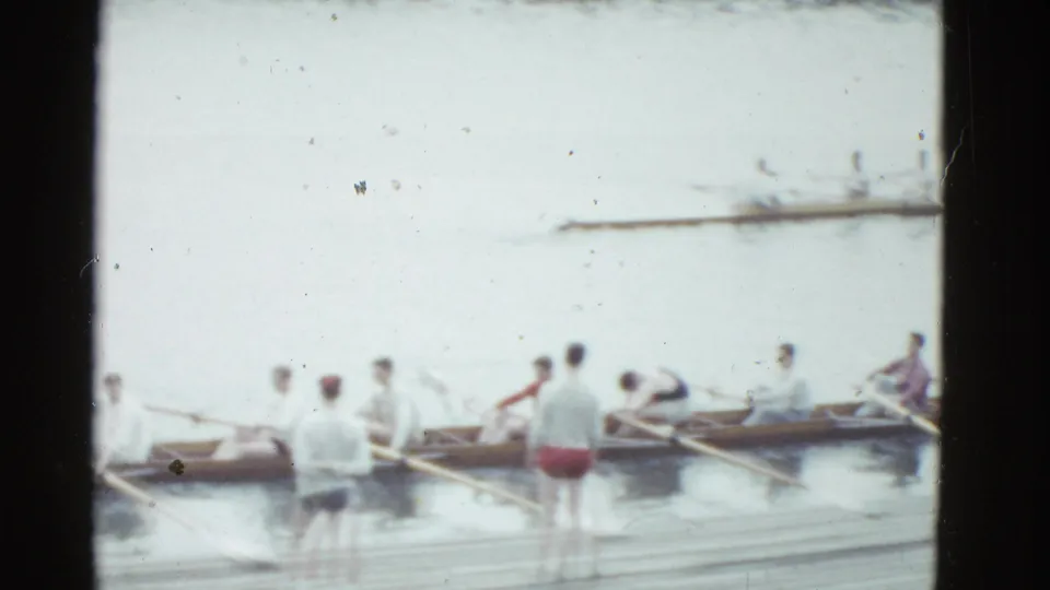 1975 Boston 8mm frame showing rowers in a long shell near a wooden dock, with standing figures watching or assisting.
