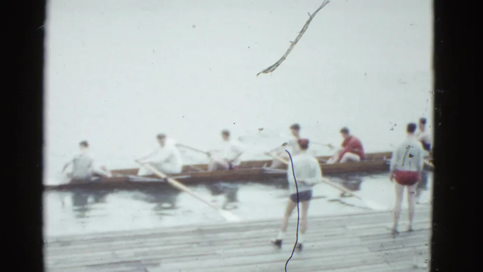 1975 Boston home-movie frame of a rowing crew aligned in a shell near the dock, with a standing figure at the right.