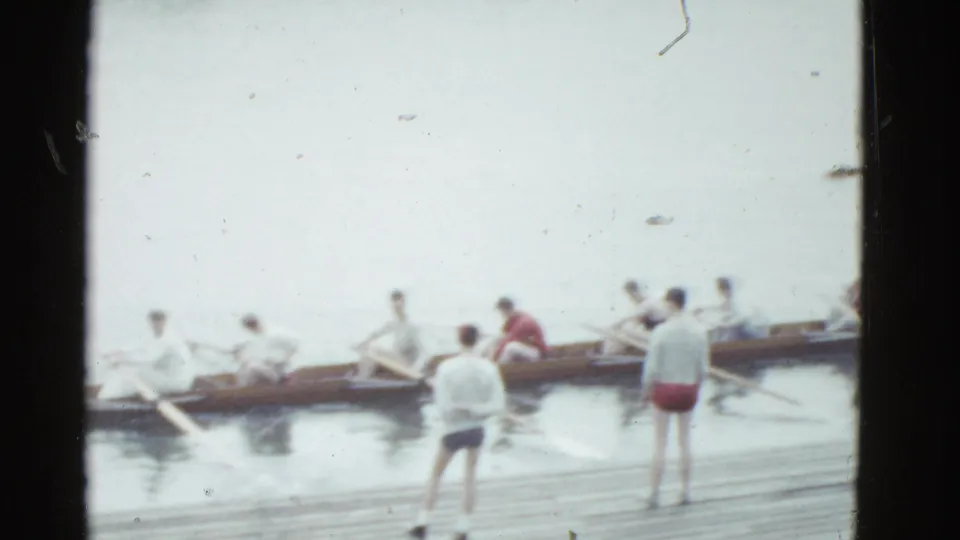 1975 Boston 8mm frame showing the rowing crew moving from the dock while two figures in shorts stand nearby.