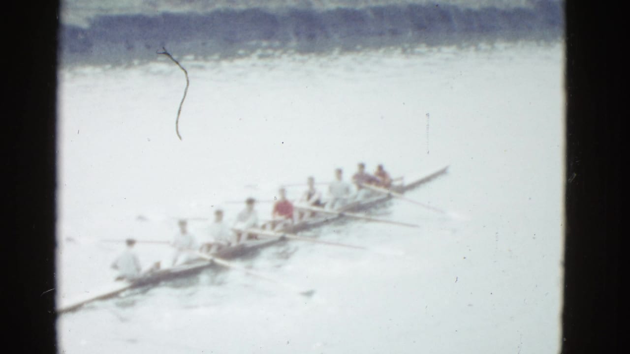 Rowing crew in a long narrow boat on a pale river, seen through an 8mm frame.