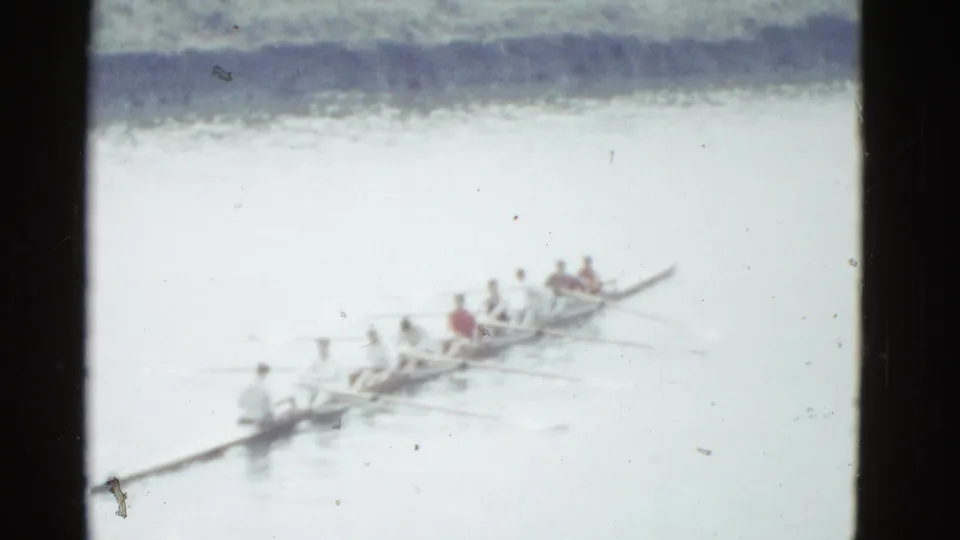 The rowing boat continues across the river, with the crew seated in line and a dark shoreline band in the background.