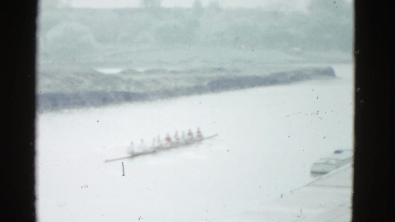 Distant rowing crew in a shell on pale river water with shoreline behind.