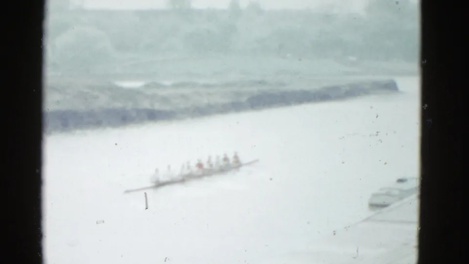 1975 Boston home-movie frame showing a distant rowing shell on a river, with low shoreline, trees, and dark 8mm frame edges.