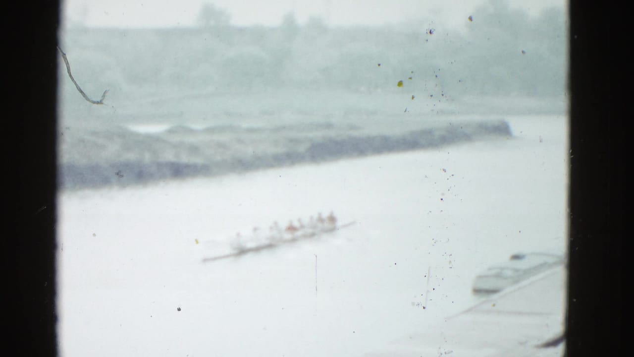 Distant 1975 Boston river view with rowers in a shell, a shoreline in the background, and a small docked boat at frame edge.