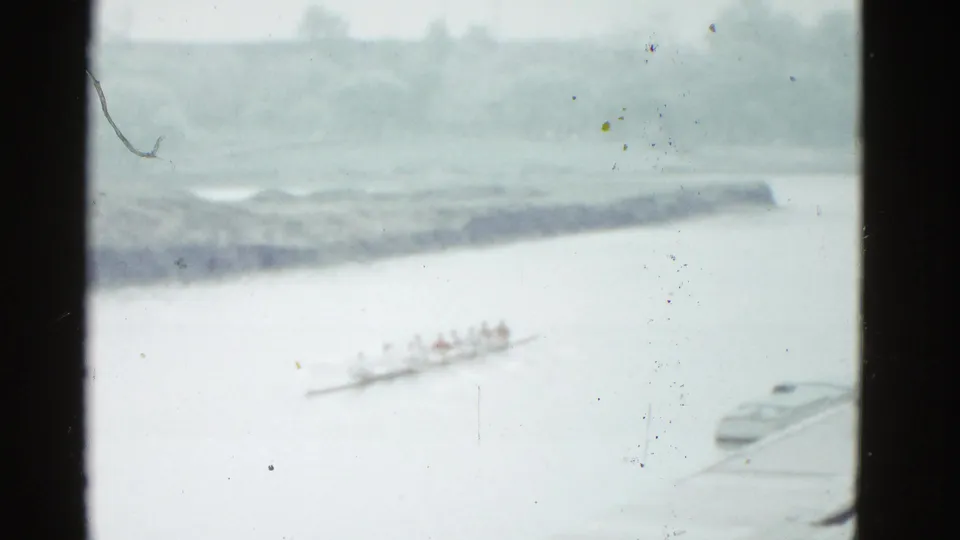 Distant 1975 Boston river view with rowers in a shell, a shoreline in the background, and a small docked boat at frame edge.