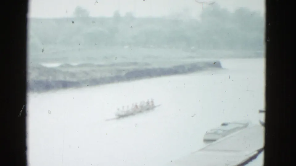 Soft 8mm frame from Boston in 1975 showing a distant rowing crew on pale water beneath a hazy tree-lined bank.