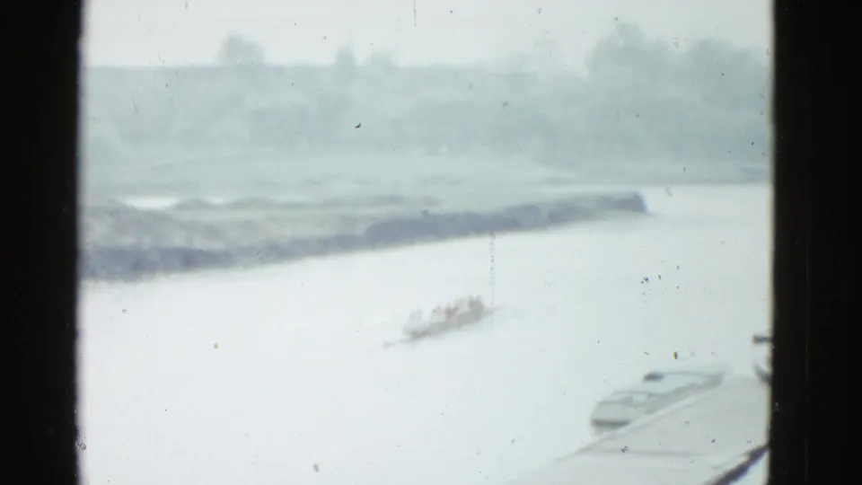 Distant 1975 Boston rowing scene with a crew shell on the river, a low shore beyond, and a small boat at the lower right.