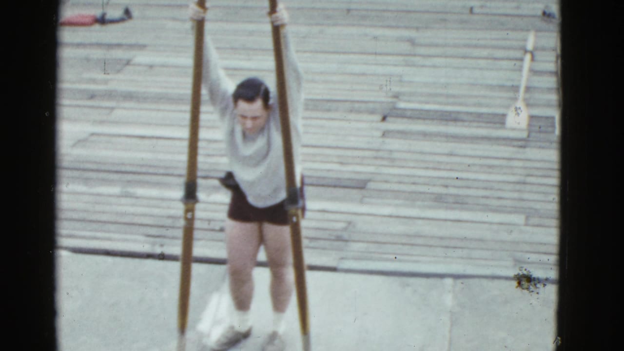 In 1975 Boston home-movie footage, a man in a light shirt and dark shorts grips tall poles on a wooden dock near water.