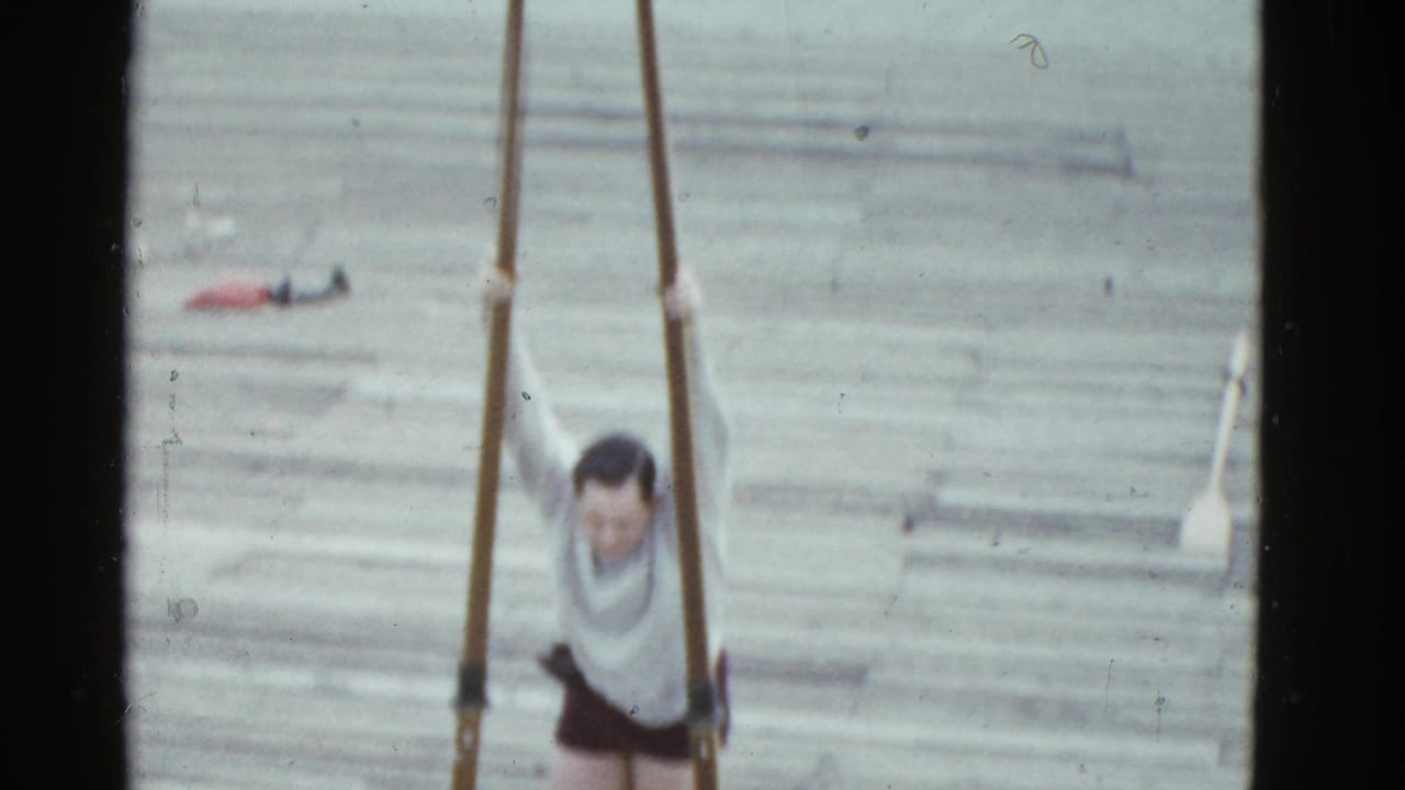 The man appears suspended between two poles on a dock, with water, boards, and a small distant boat visible behind him.