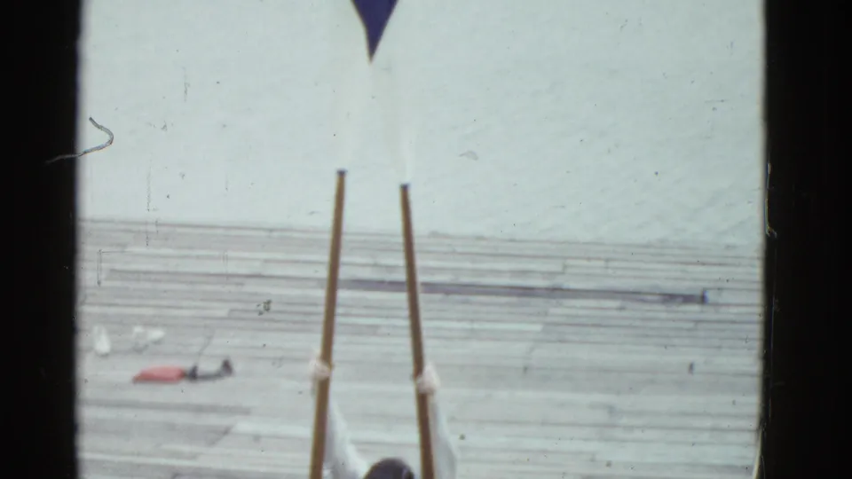 1975 Boston 8mm frame showing rowing oars upright on a wooden dock with part of a man entering at the bottom edge.