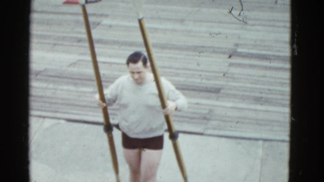 1975 Boston 8mm frame of an adult man carrying long oars across a wooden dock before rowing.