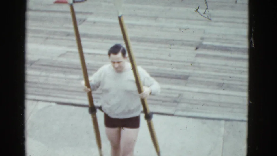 1975 Boston 8mm frame of an adult man carrying long oars across a wooden dock before rowing.
