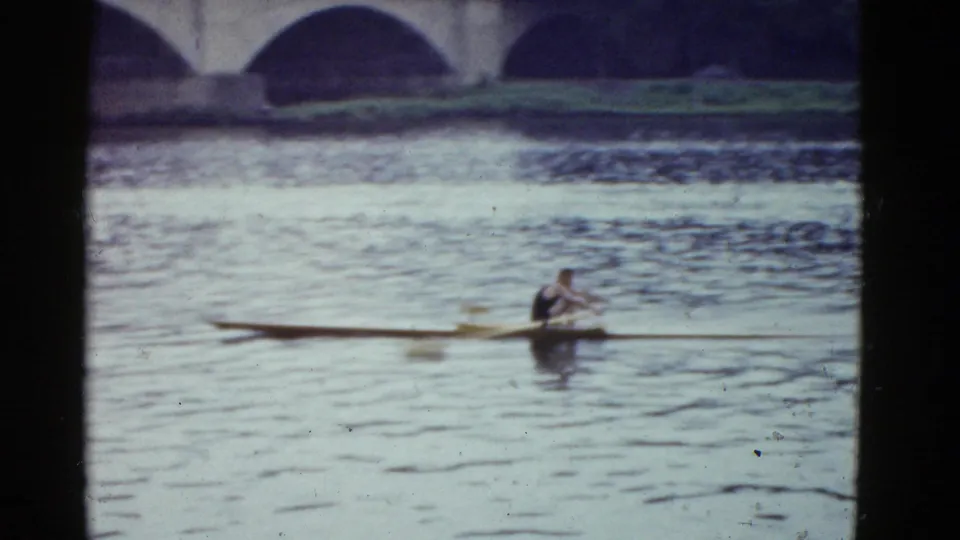 1975 Boston 8mm frame showing a single sculler on the river with arched bridge spans behind him.