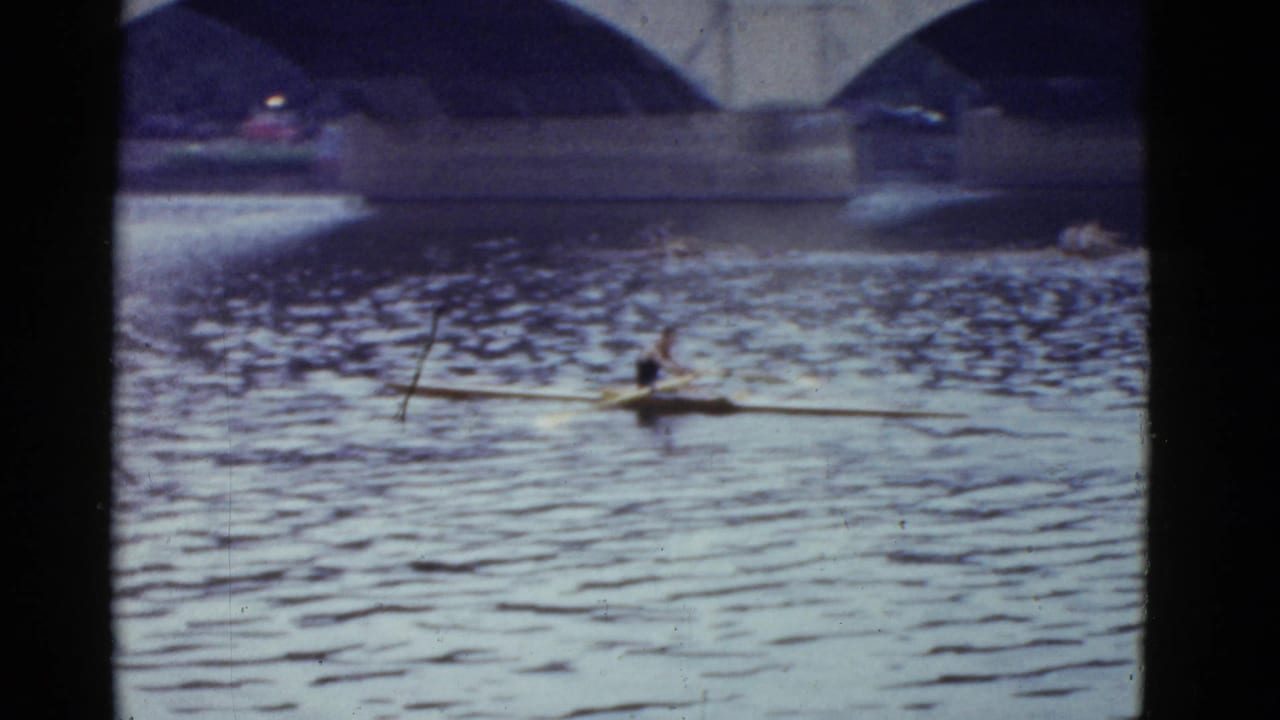 1975 Boston home-movie frame of a single scull rower on rippled water with bridge arches and distant boats behind him.