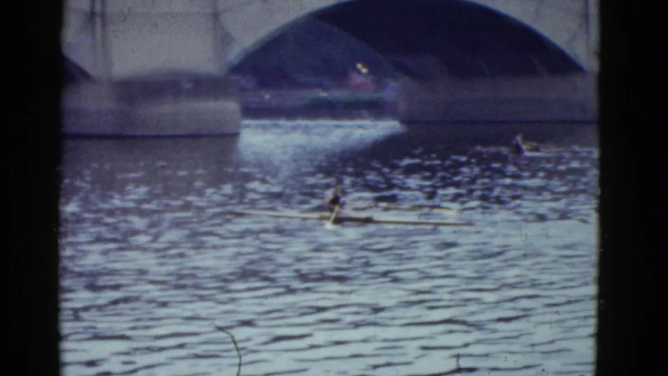 1975 Boston 8mm frame with the sculler farther along the water, framed by bridge arches and soft home-movie focus.
